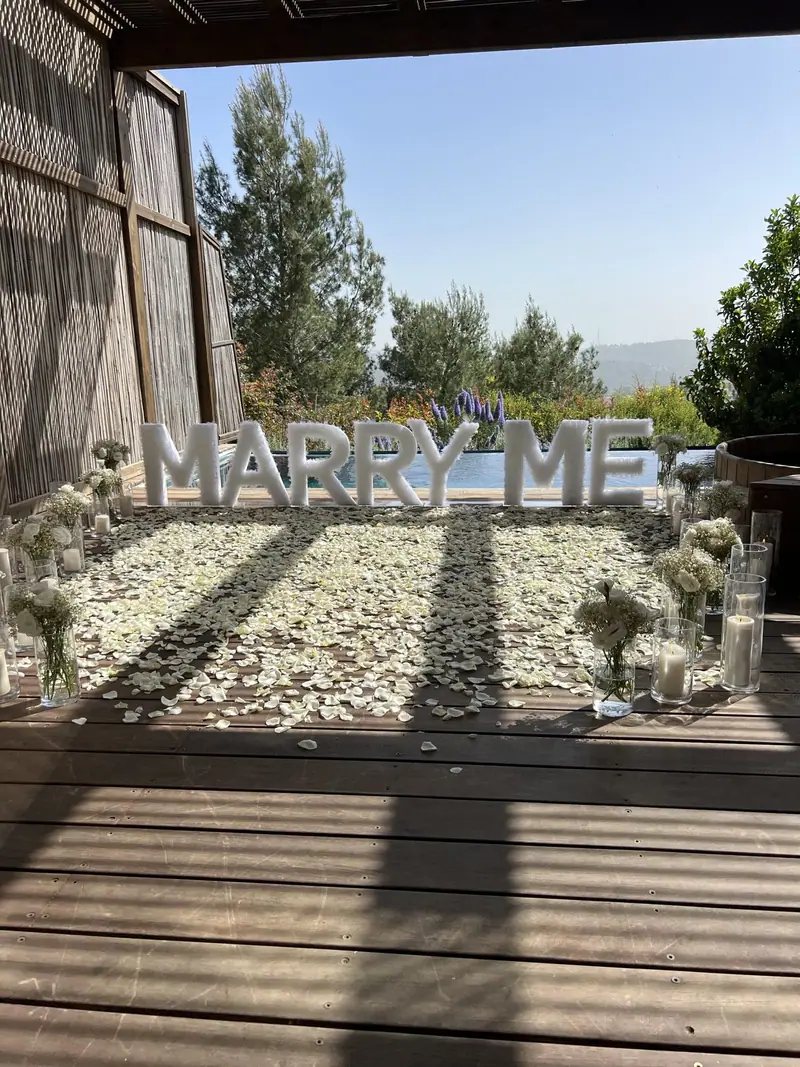 אותיות MARRY ME לבנות וגדולות מעל רצפת עלי כותרת לבנים על דק עץ