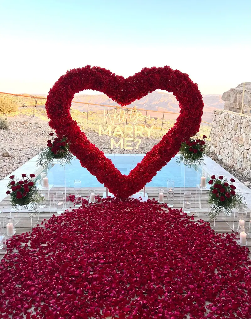 לב ורדים אדומים עם שלט ניאון מעל שטיח עלי ורדים אדומים, ליד בריכה עם נוף לים