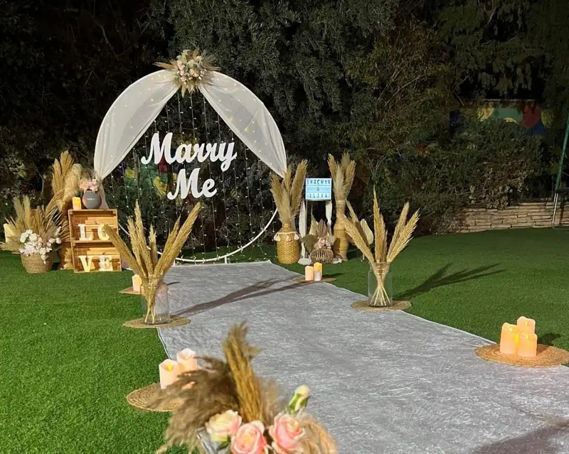 קשת בד לבנה עם שלט Marry Me, פמפס וארגזי עץ בקצה שביל שטיח אפור על דשא בערב